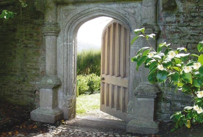 Old granite stone archway with open wooden door leading to a garden, sunlight filtering through.